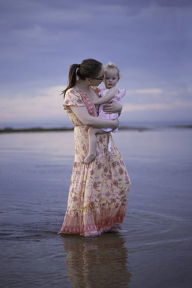 A woman holding a young girl while standing in shallow water at the beach during sunset, both dressed in pink and floral clothing.