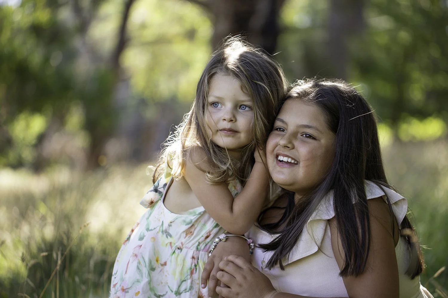 Two young girls outdoors in a grassy area, one with blue eyes and light brown hair, the other with dark brown hair, smiling and hugging each other.