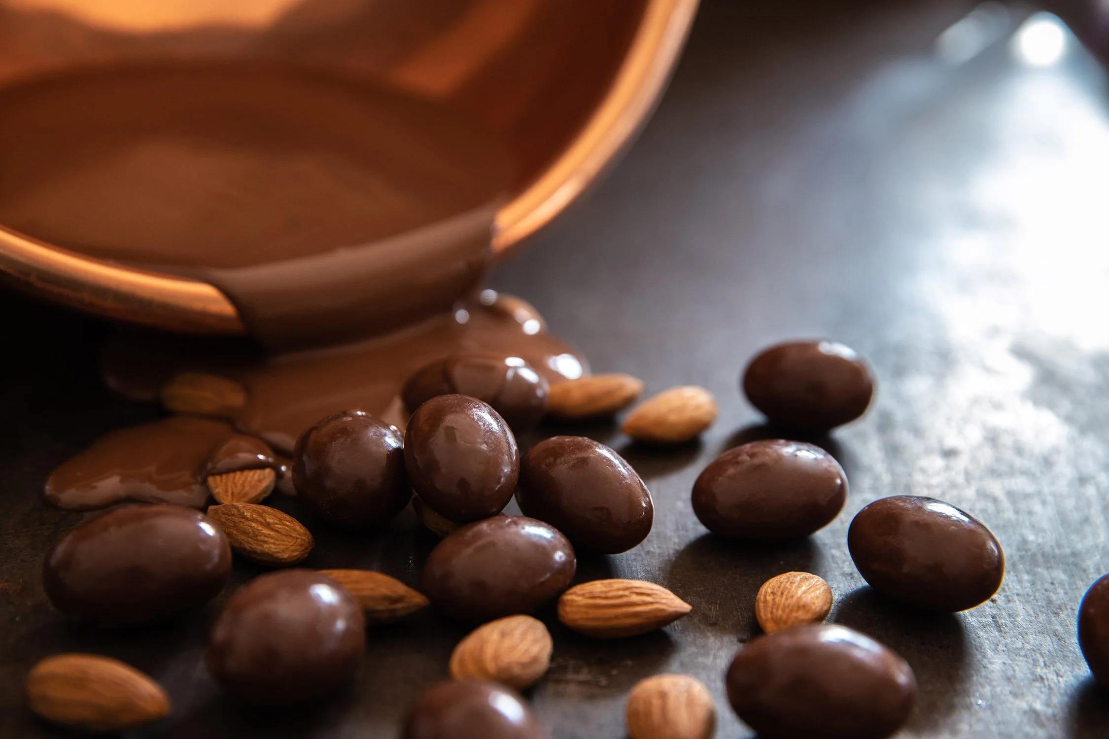 Chocolate and almonds spilled from a copper bowl onto a dark surface.