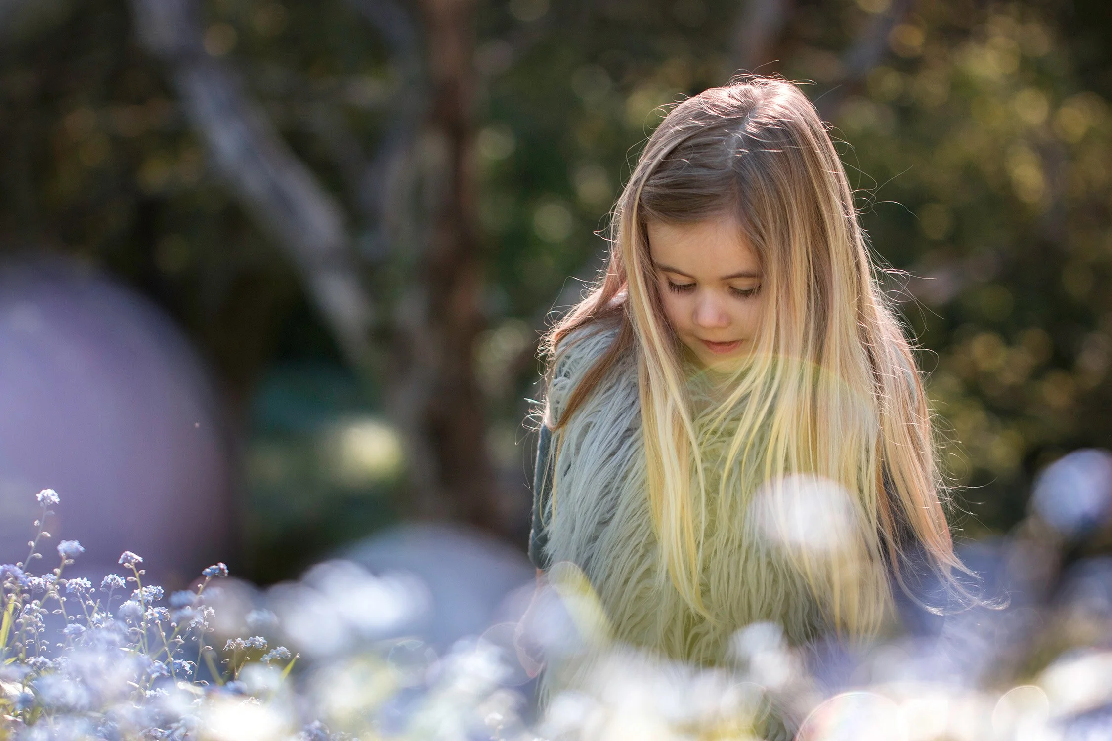 A young girl with long blonde hair looking down outdoors surrounded by blurred white flowers and greenery in the background.