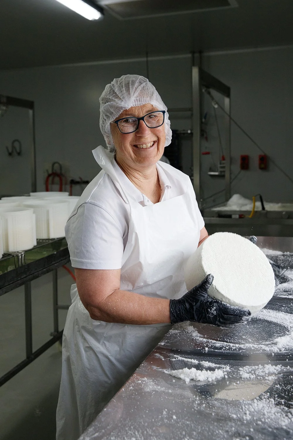 A smiling woman in a hairnet, glasses, and white uniform holding a large wheel of cheese in a cheese-making facility