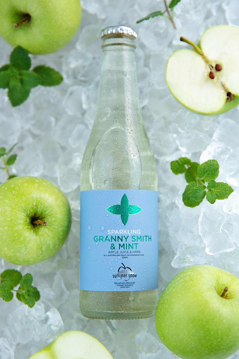 A clear glass bottle of sparkling Granny Smith & Mint apple juice on ice, surrounded by whole and sliced green apples and mint leaves.