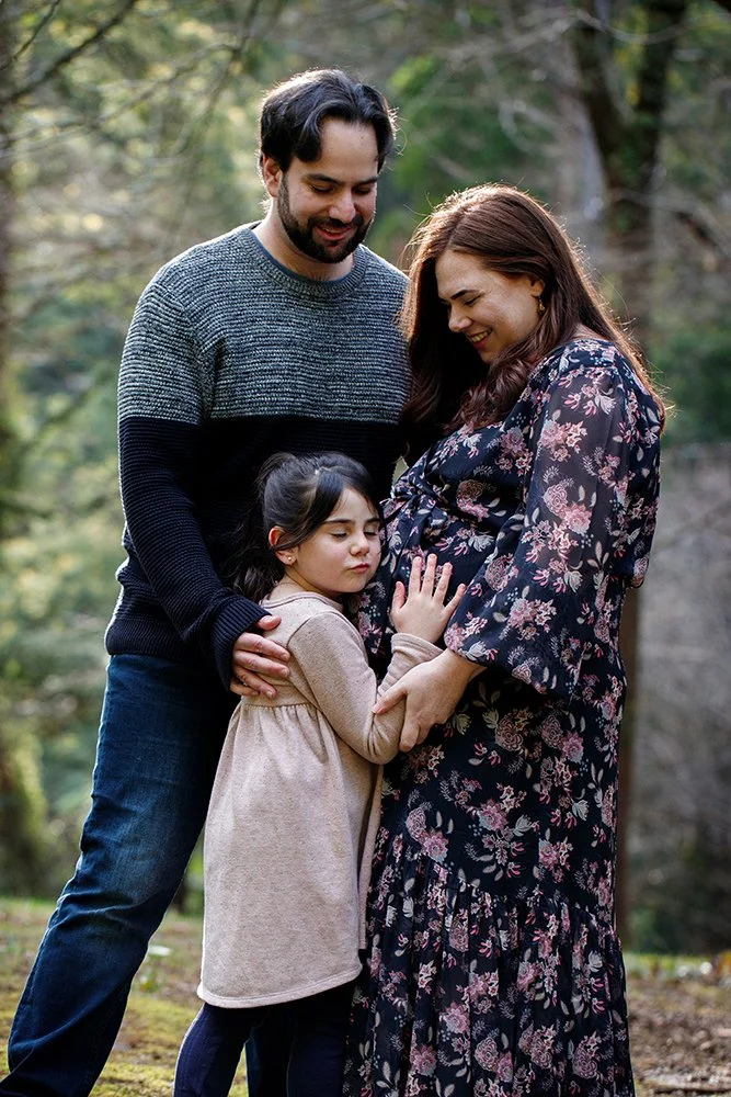 A family of three with a pregnant woman, a man, and a young girl standing outdoors in a wooded area, sharing an emotional moment.