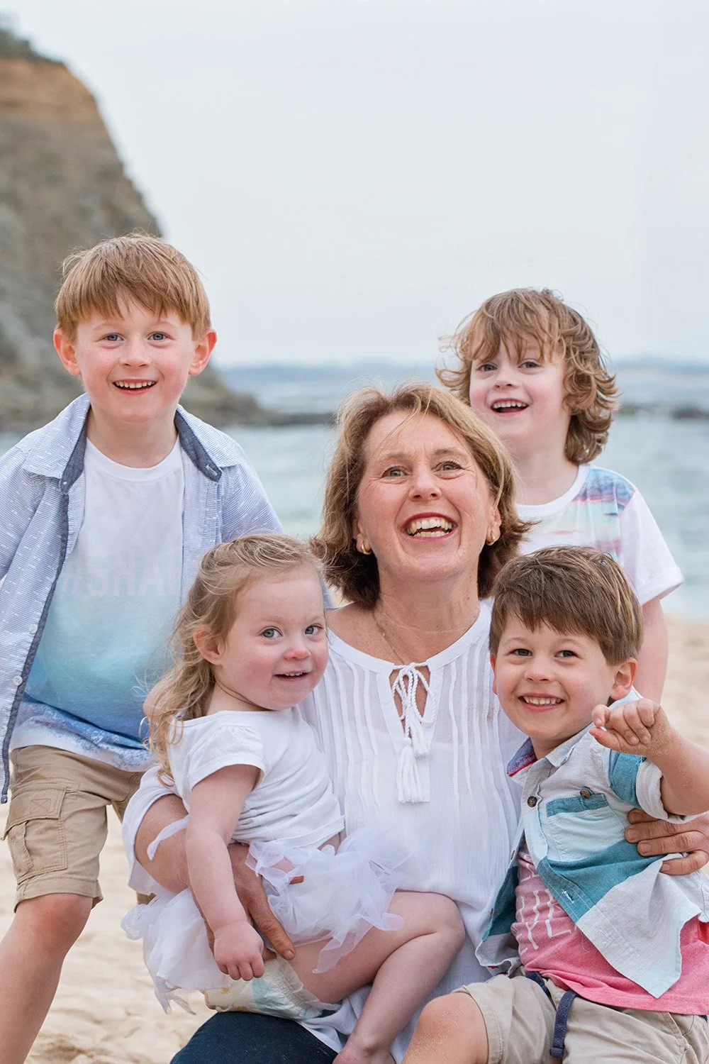 A smiling woman holding a young girl on her lap at the beach, surrounded by three boys, with cliffs and ocean in the background.