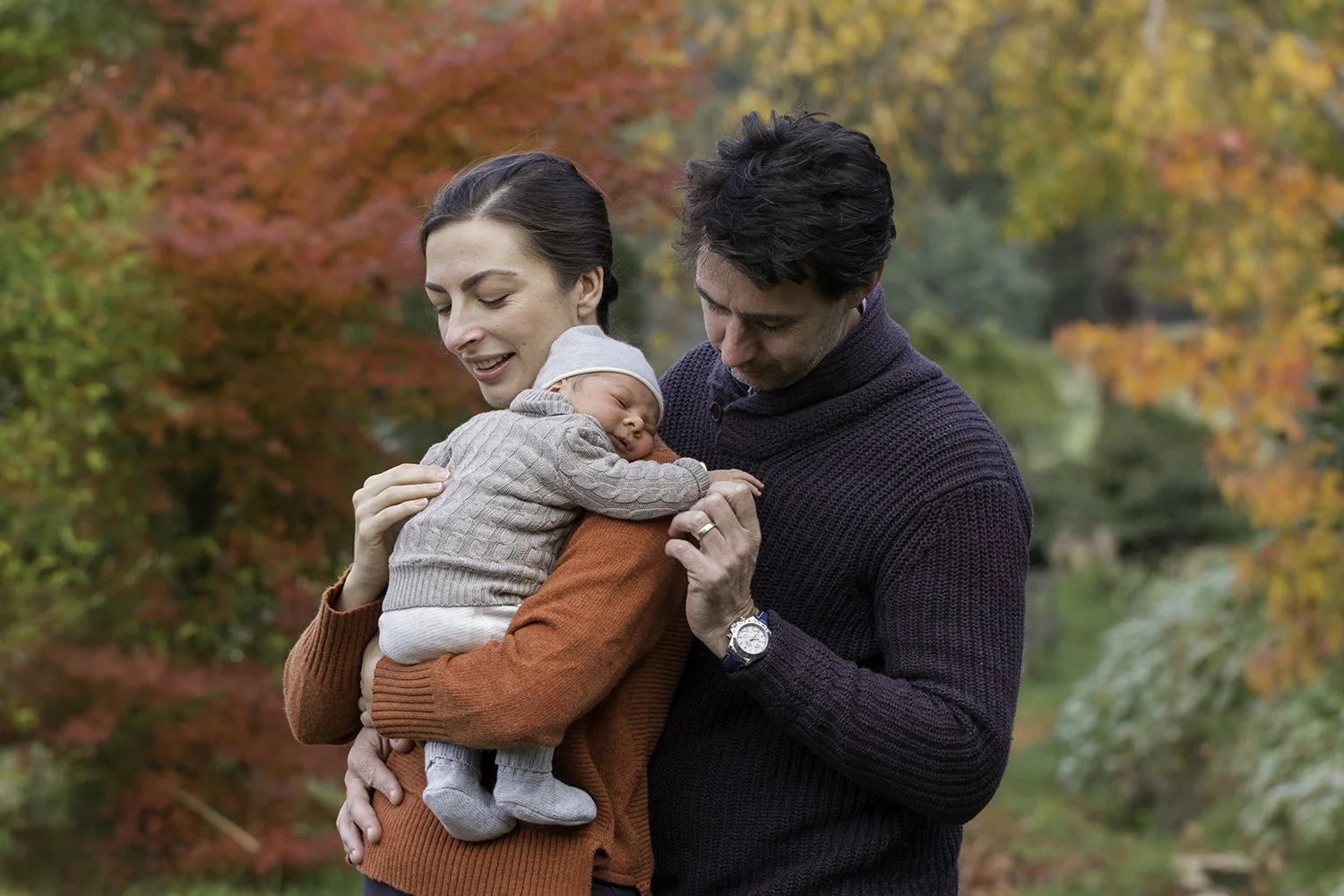 A family of three, a woman, a man, and a sleeping baby, outdoors in a park with autumn foliage in the background.