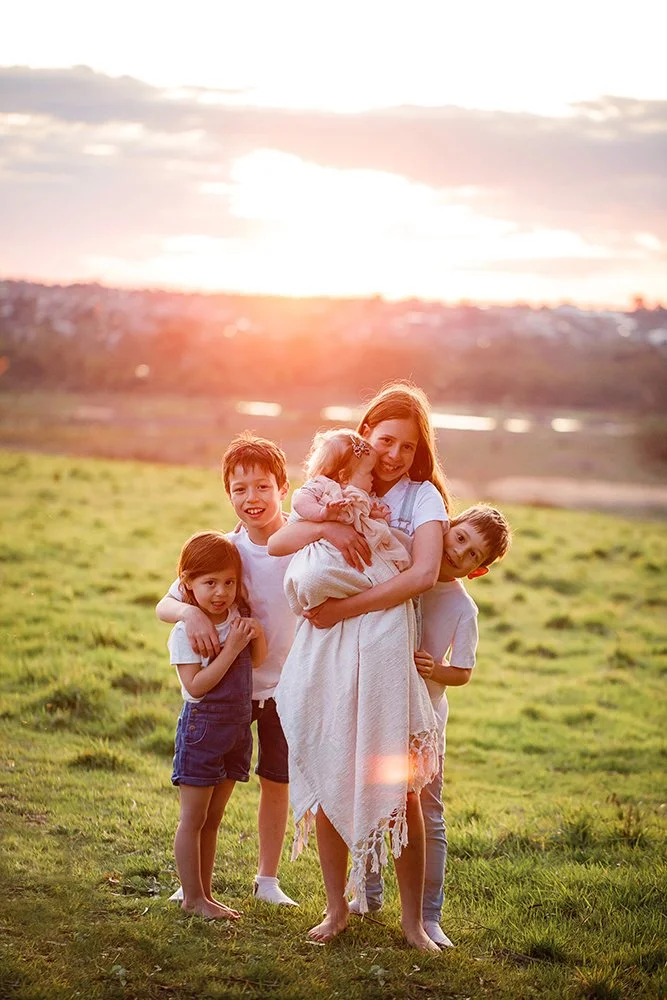 A woman holding a young girl in a white dress, standing outdoors on a grassy field at sunset with four children, smiling and embracing each other.
