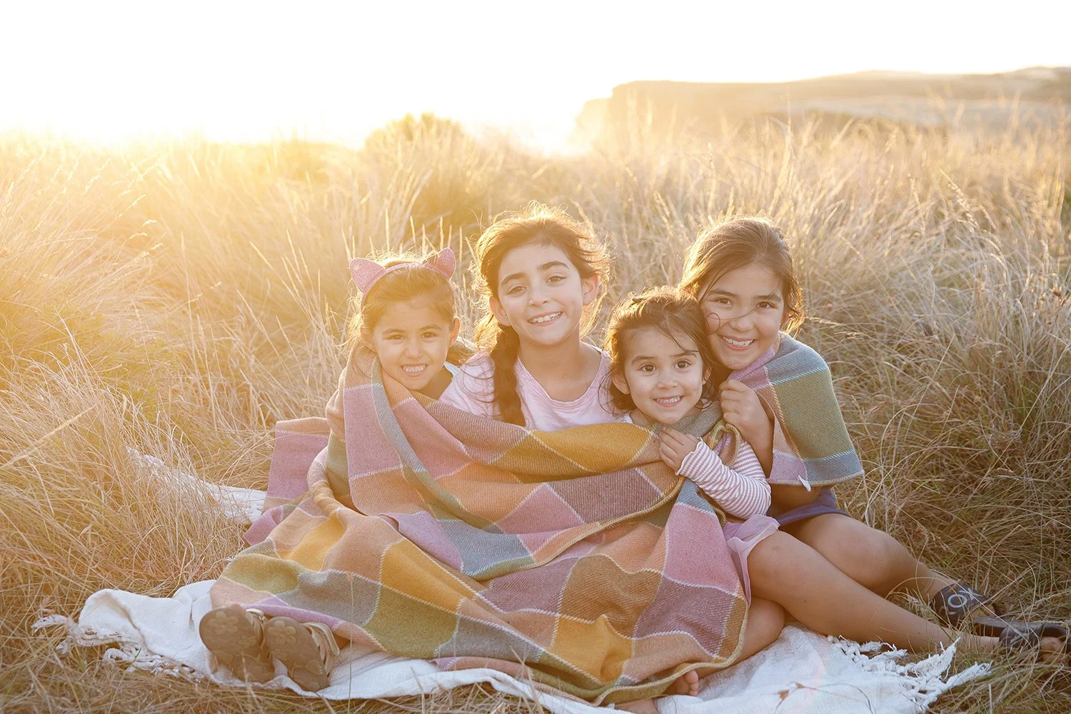 Five children sitting together on a blanket in a field of tall grass during sunset, wrapped in a colorful blanket, smiling at the camera.