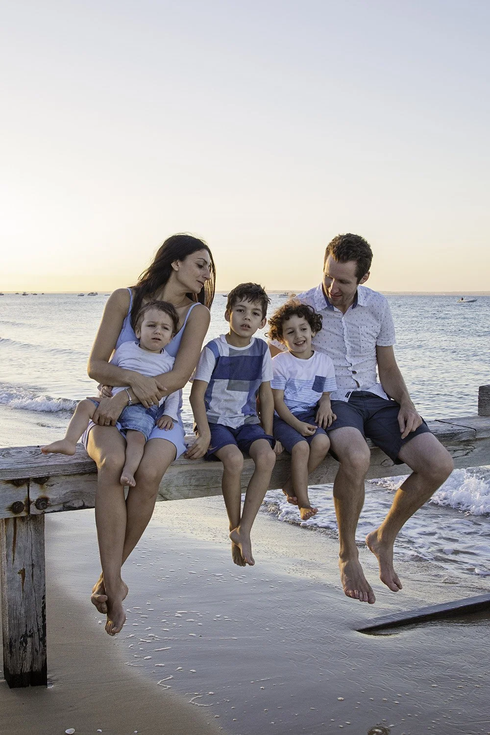 A family of five sitting on a wooden dock at the beach during sunset, with the ocean in the background. The parents are seated with their three children, all looking relaxed and enjoying the moment.