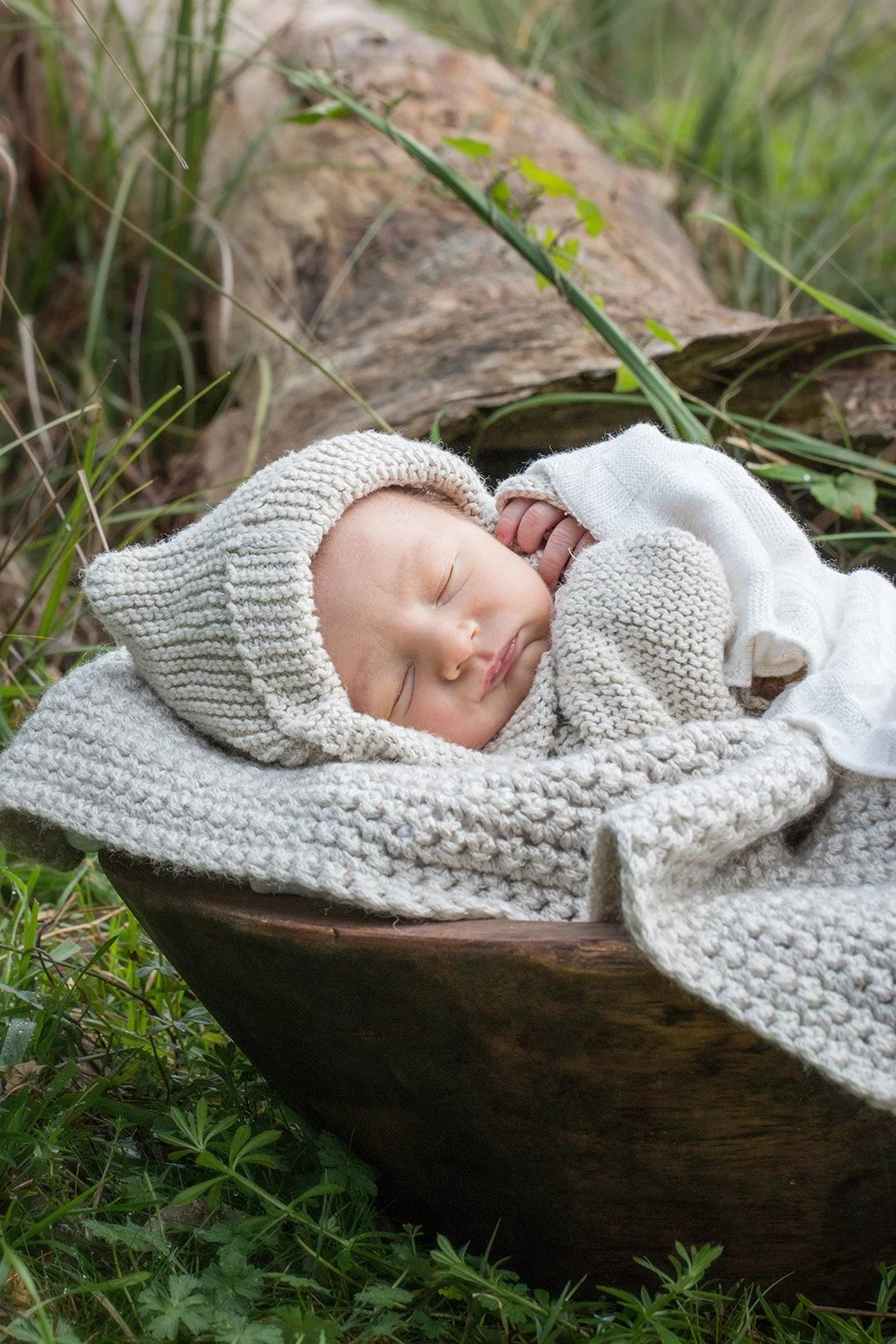 A sleeping baby with a knitted hat and blanket, lying in a wooden bowl outdoors among green grass and plants.