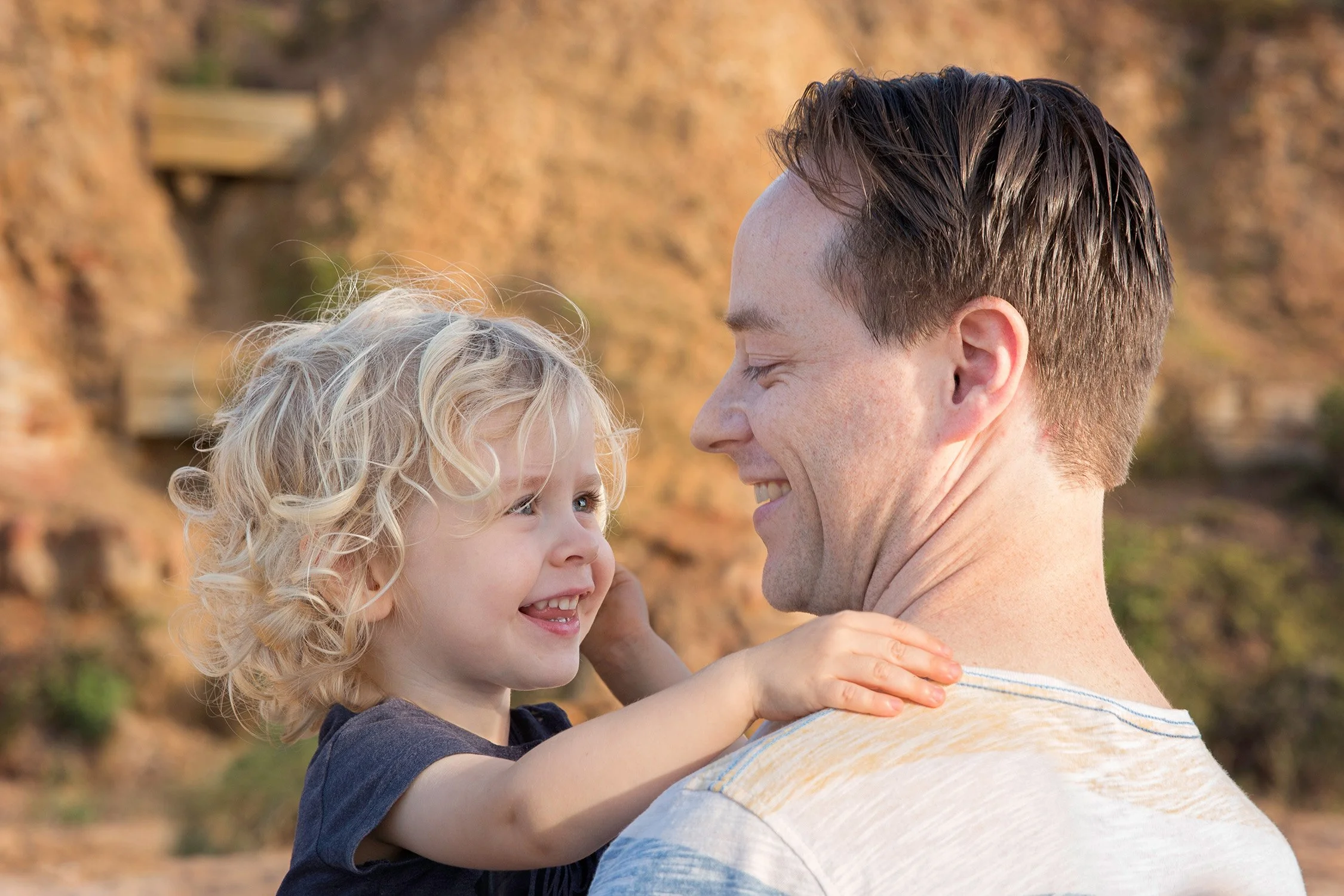 A father and his young daughter smiling and looking at each other outdoors with a rocky background.