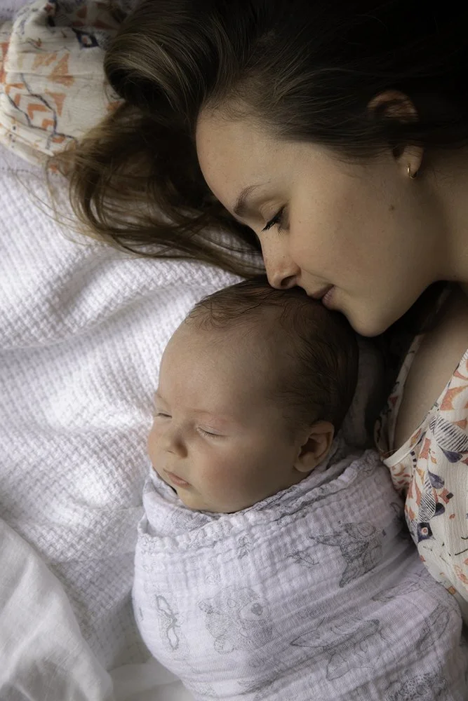 A mother with brown hair lying next to her sleeping baby wrapped in a white blanket, both resting on a bed.