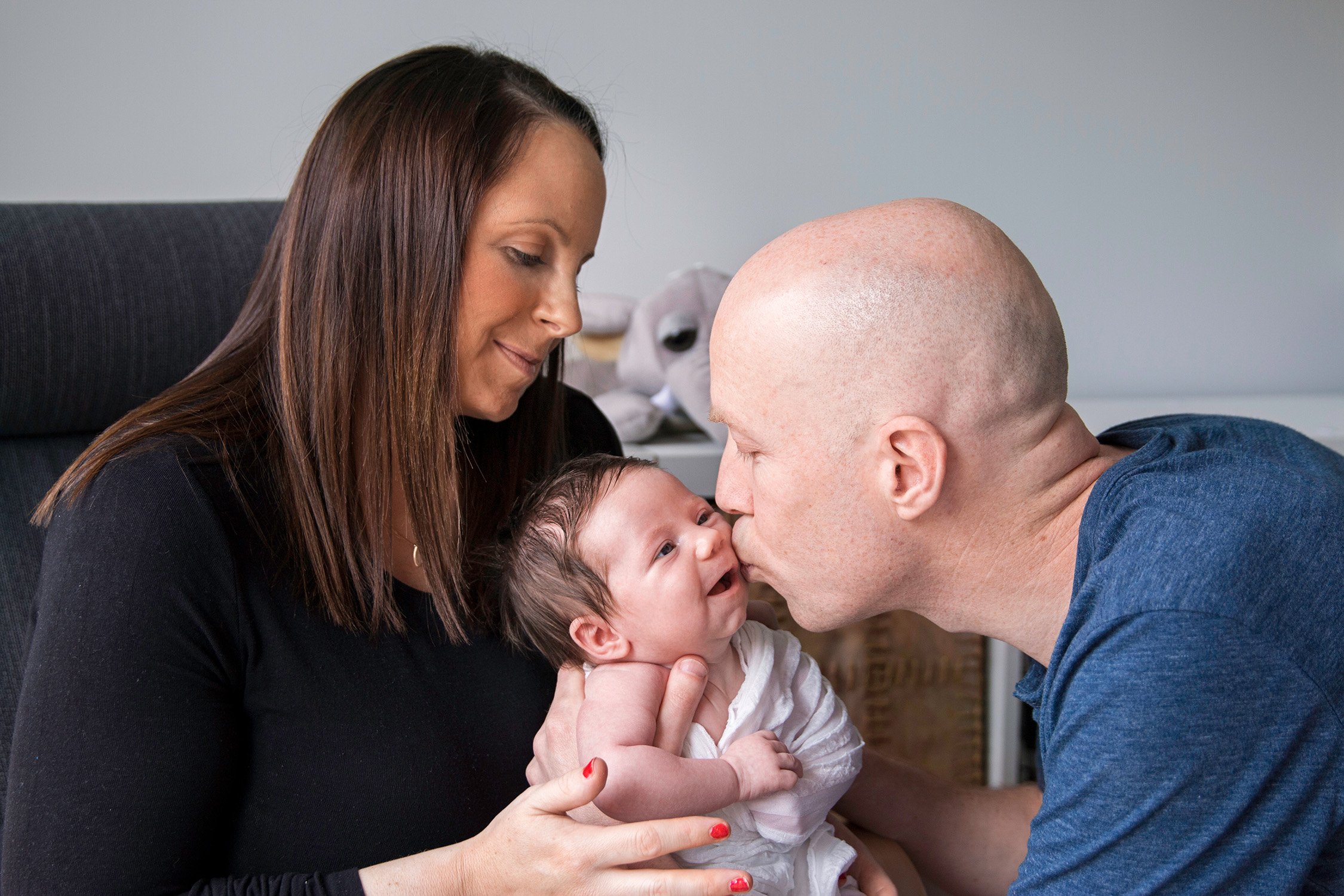 Bald man kissing a baby girl with a woman holding the baby in a room.