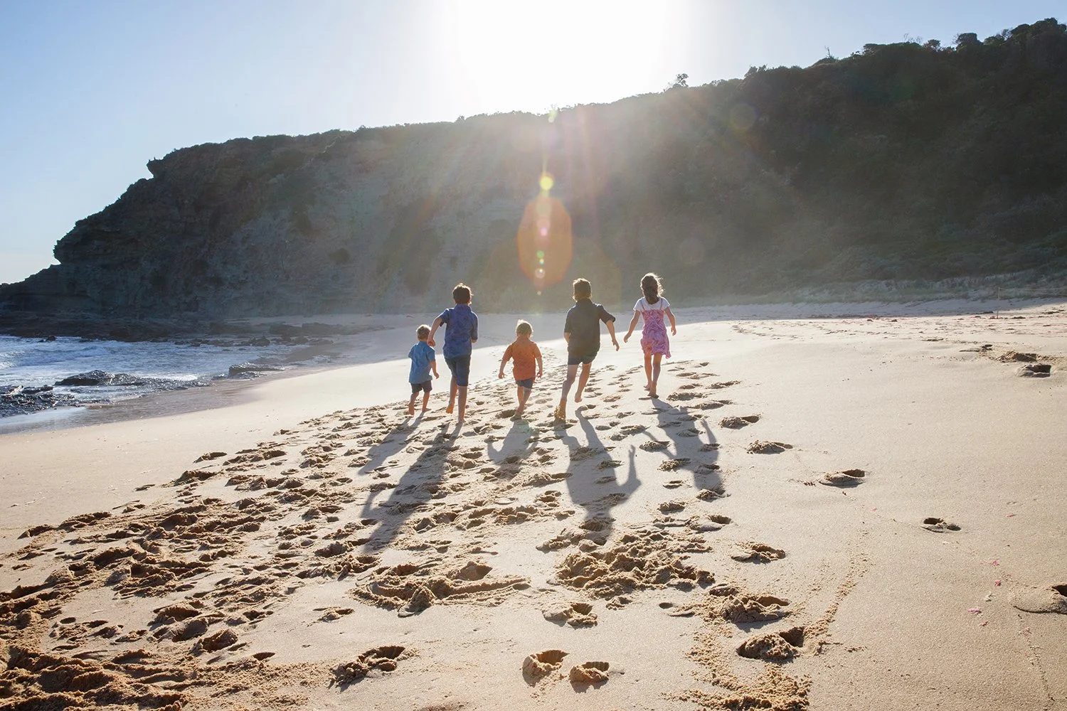 Five children running along a sandy beach during sunset, with footprints trailing behind them and a large hill in the background.
