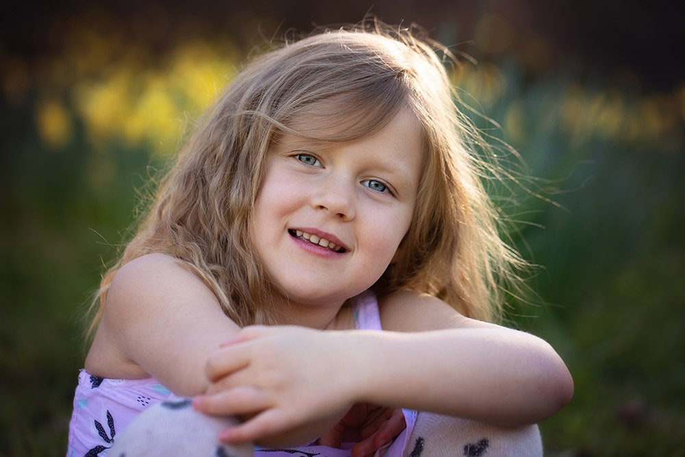 A young girl with blond, curly hair sitting outdoors with her arms crossed on her knees, smiling at the camera with a blurred green and yellow background.