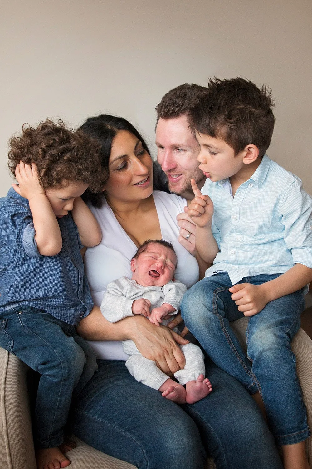 Family of five, including two children, two adults, and a newborn baby, sitting together on a beige chair against a plain wall.