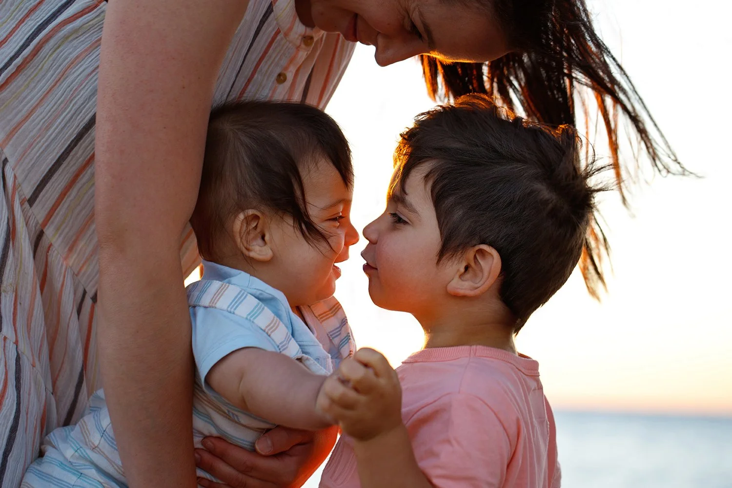 A woman with red hair leans over two children, one boy and one girl, face-to-face at the beach during sunset, with the ocean visible in the background.