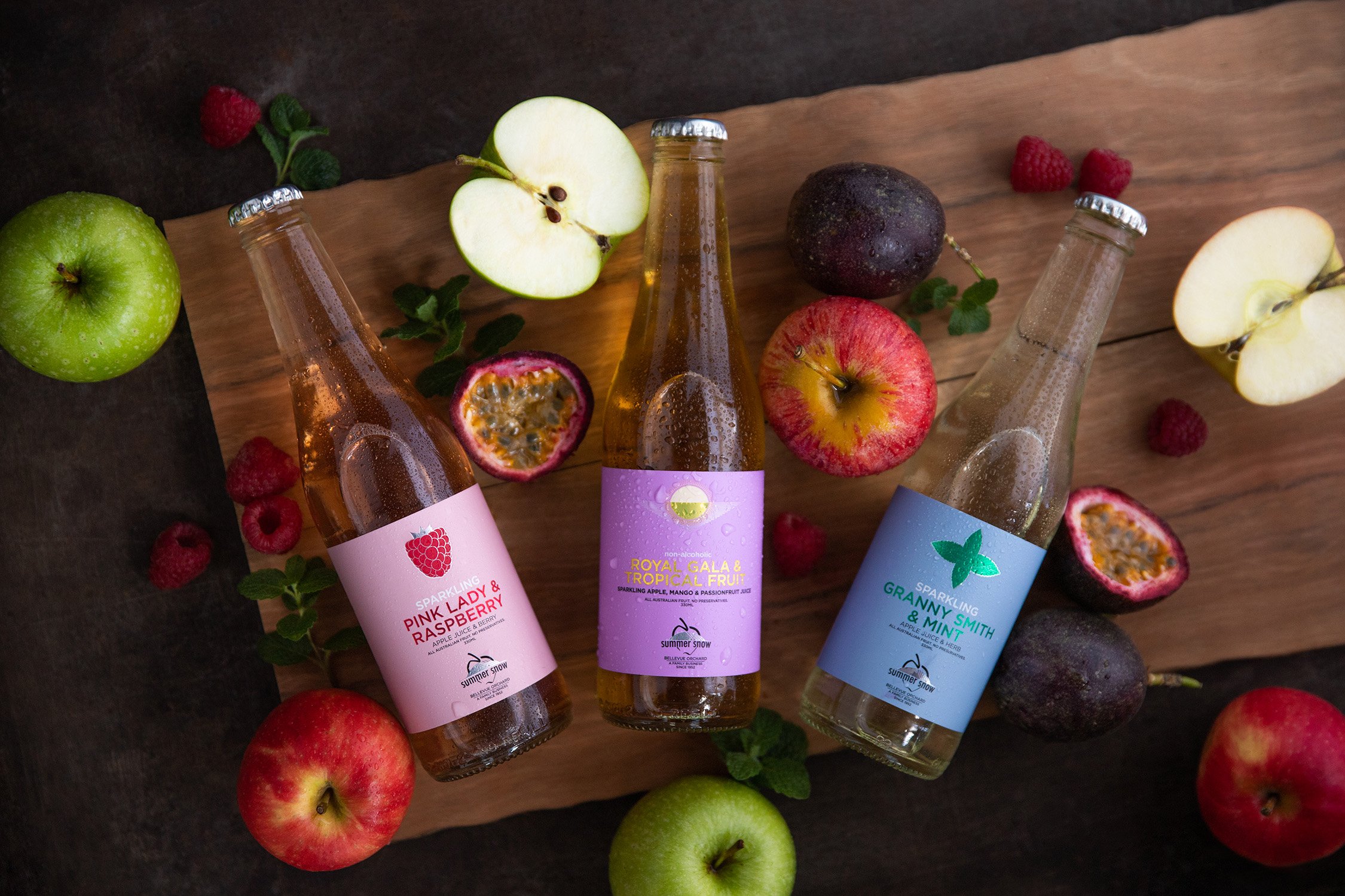 Three bottles of sparkling juice labeled Pink Lady & Raspberry, Royal Gala & Tropical Fruit, and Granny Smith & Mint, arranged on a wooden board surrounded by whole and halved apples, passion fruits, and raspberries.
