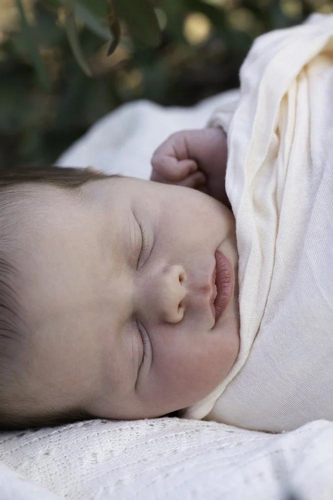 Close-up of a peacefully sleeping baby lying on a white blanket outdoors, wearing a white outfit.