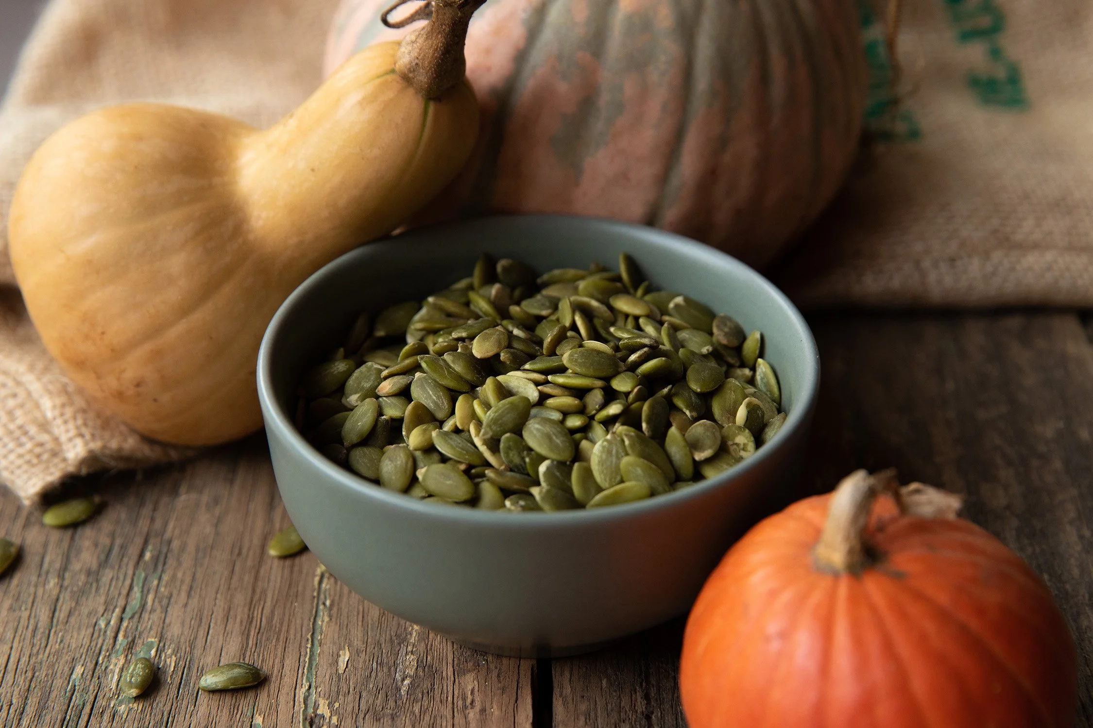 A bowl of pumpkin seeds on a rustic wooden table with whole pumpkins and a squash in the background.