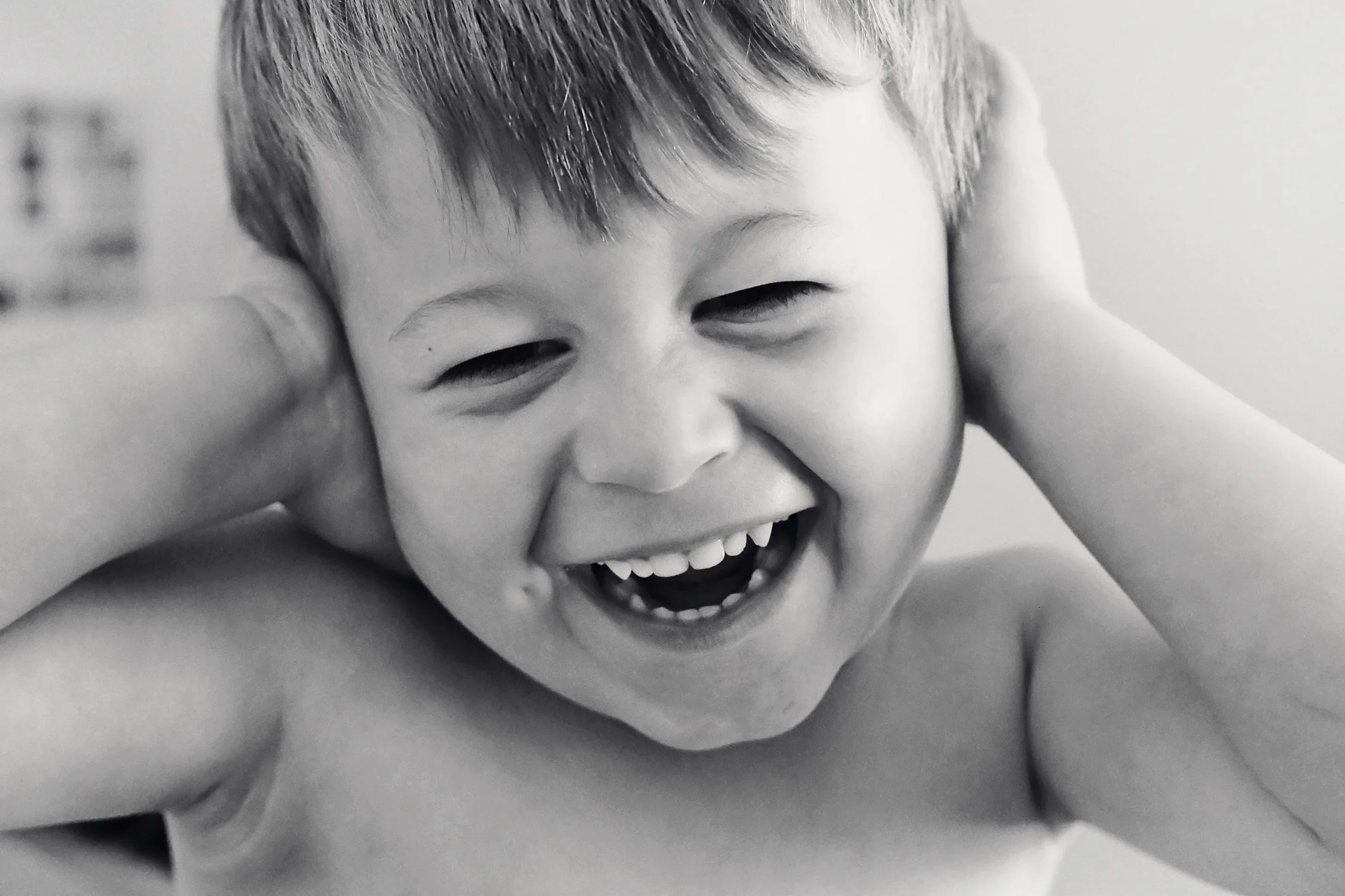 A young boy with short hair laughing with his hands covering his ears in a black and white photo.