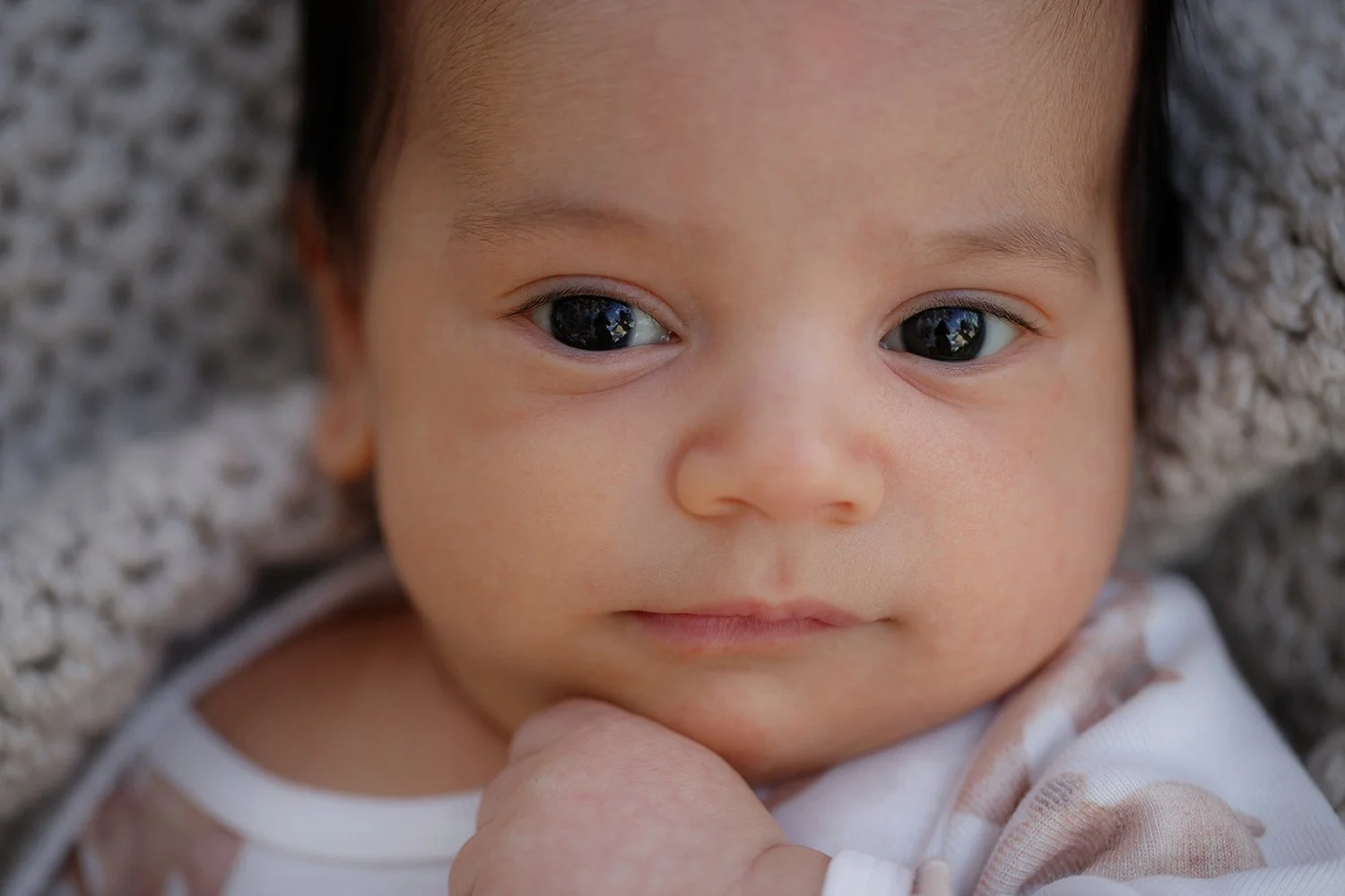 Close-up of a baby's face lying on a textured surface with hand near chin, with dark eyes and smooth skin.