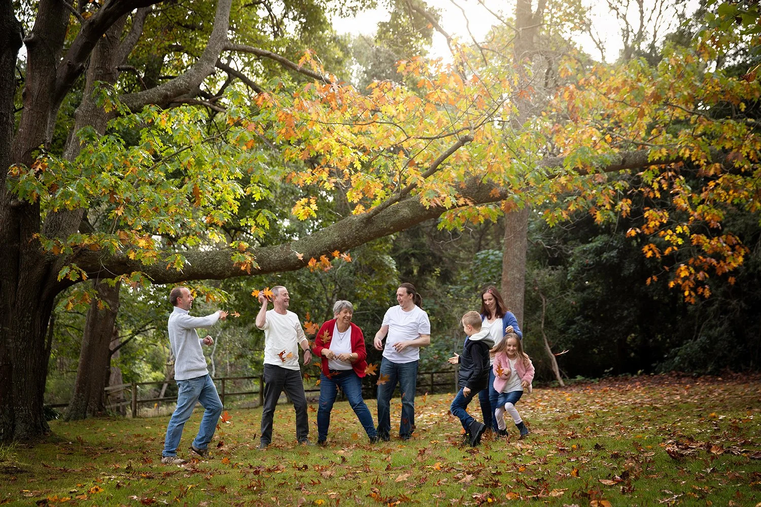 A multi-generational family playing and laughing in a park during autumn, surrounded by fallen leaves and trees with colorful fall foliage.
