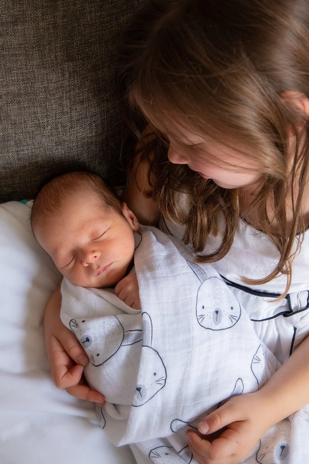 A young girl tenderly holding a sleeping baby, wrapped in a cloth with a bear face pattern, on a bed or sofa.