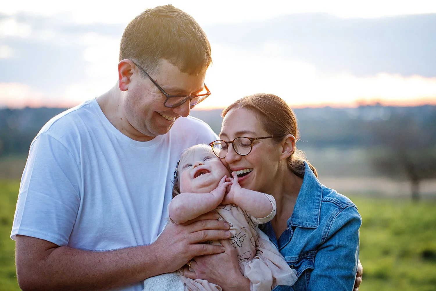 A family of three smiling outdoors at sunset. The father and mother are holding their young daughter, who is laughing and touching her face.