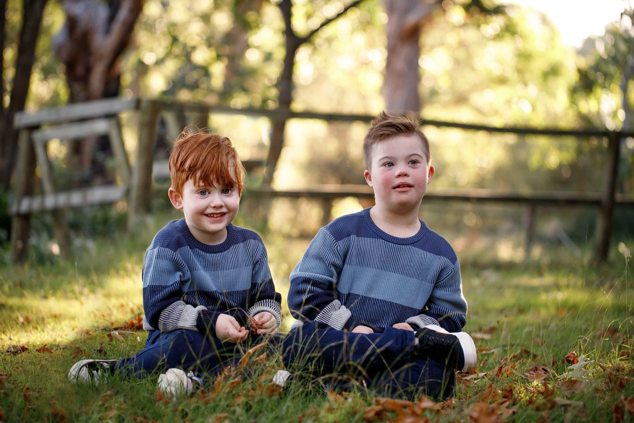Two young boys sitting on the grass in a park during autumn, surrounded by fallen leaves and trees with sunlight filtering through.
