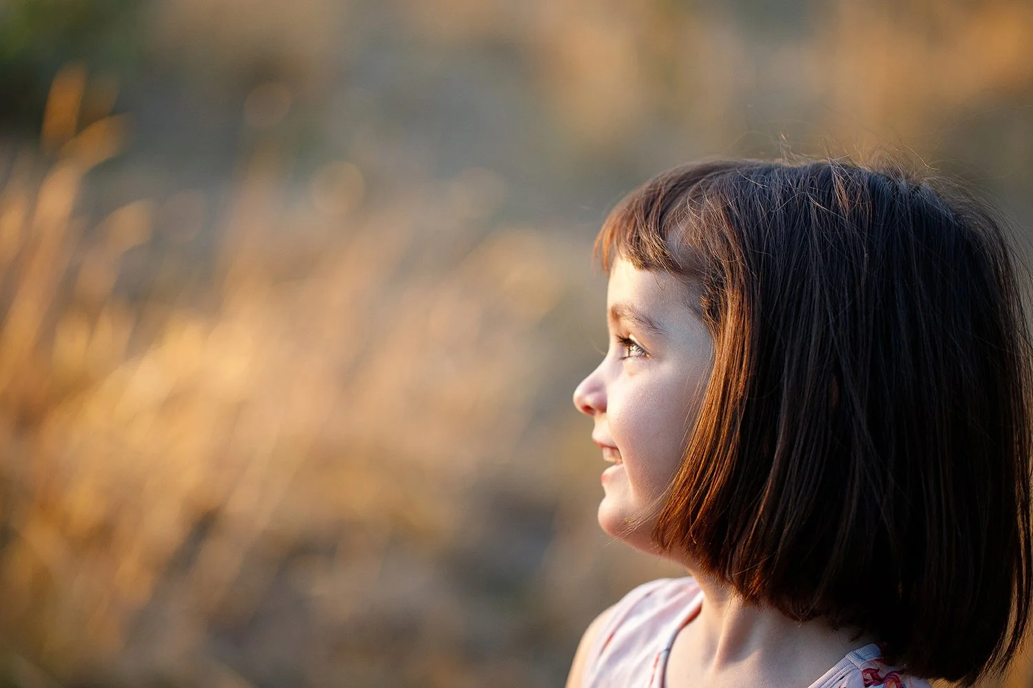 Profile of a young girl with short brown hair, smiling while looking to the right, in an outdoor setting during sunset.