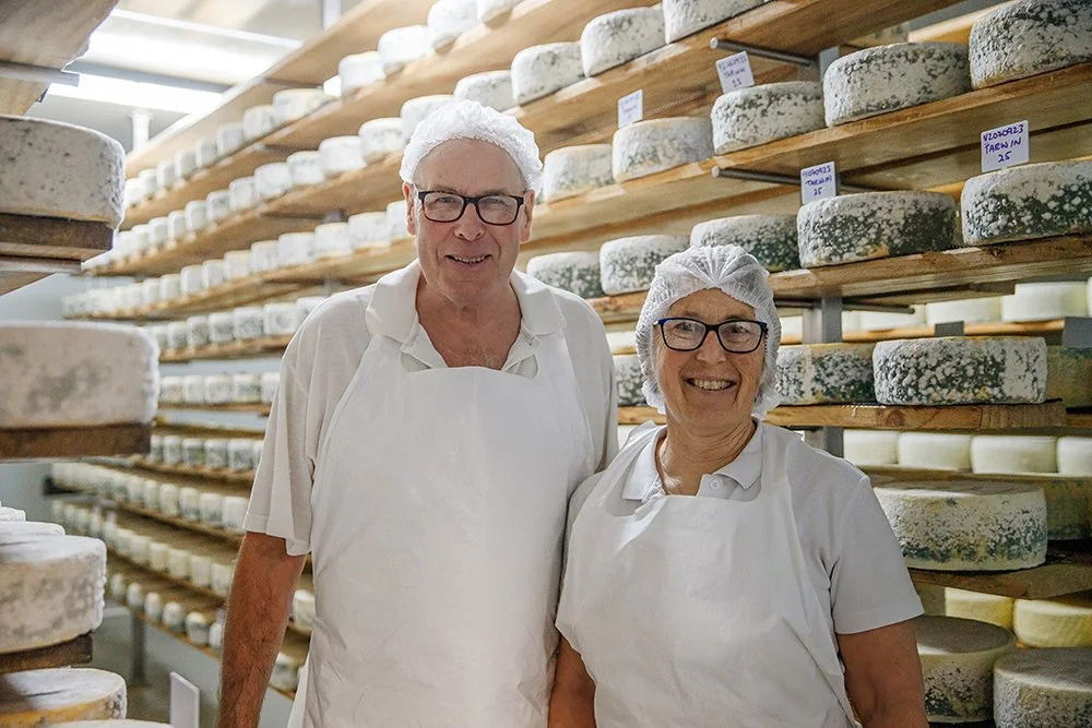 A man and a woman wearing hairnets, glasses, and white aprons in a cheese aging room surrounded by racks of large cheese wheels.