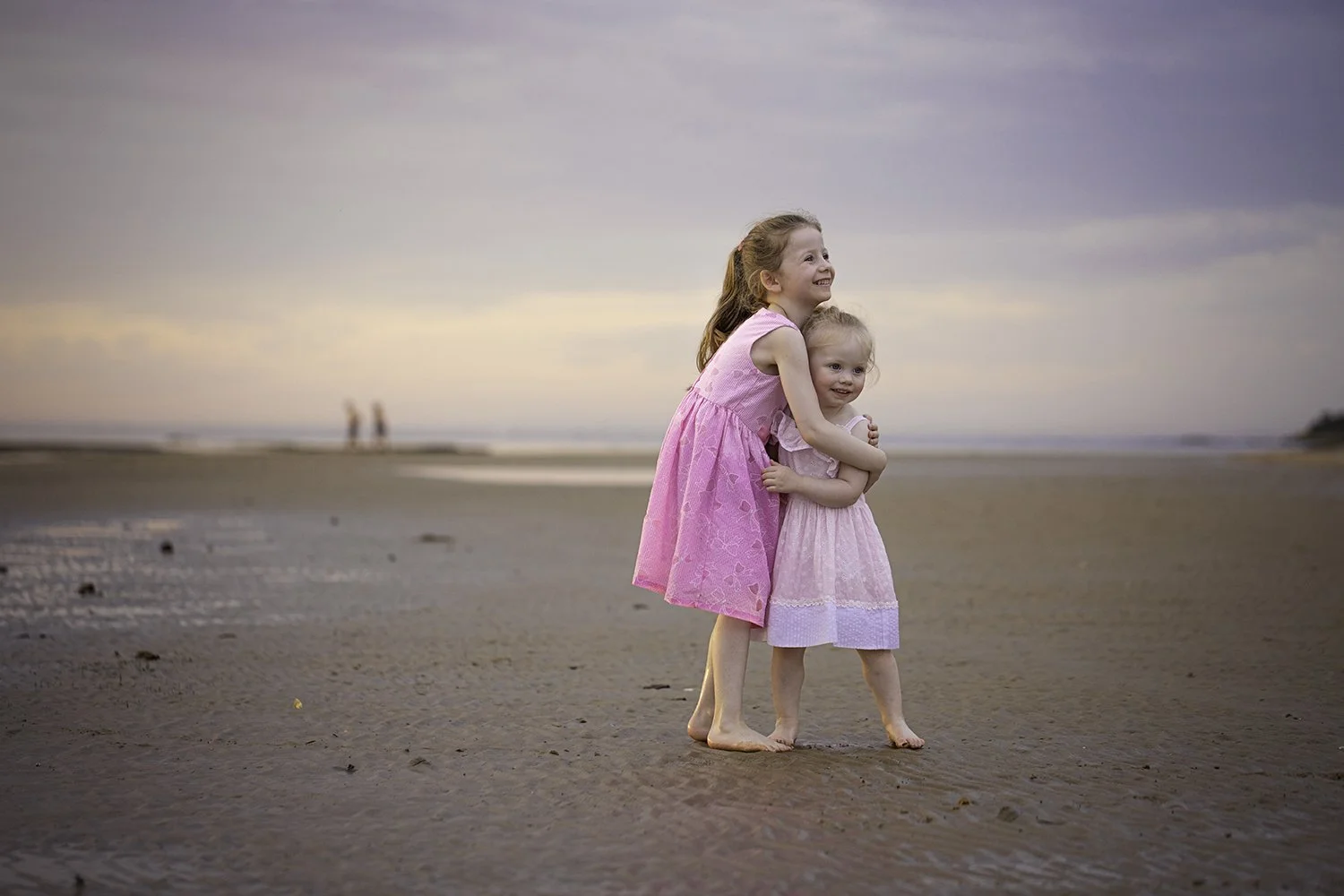 Two young girls hugging on a beach at sunset, wearing pink dresses, with water and distant horizon in the background.
