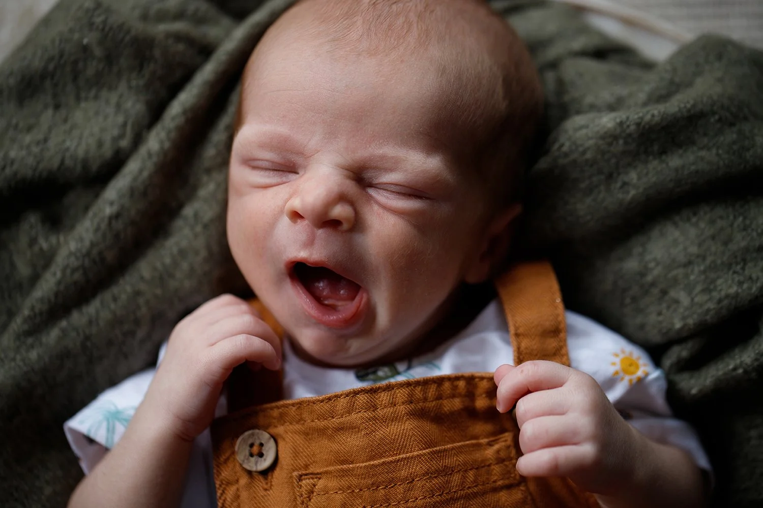 Close-up of a yawning baby with closed eyes, wearing a white shirt with colorful designs and orange overalls, lying on a soft textured blanket.