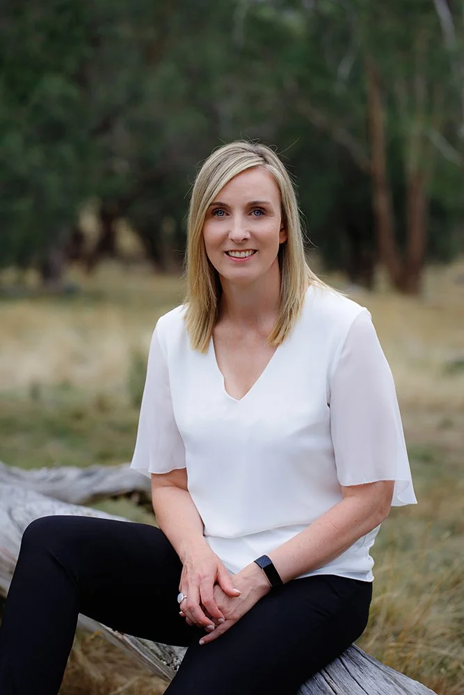 A woman with shoulder-length blonde hair sitting outdoors on a log, wearing a white blouse and black pants, with a green forest background.