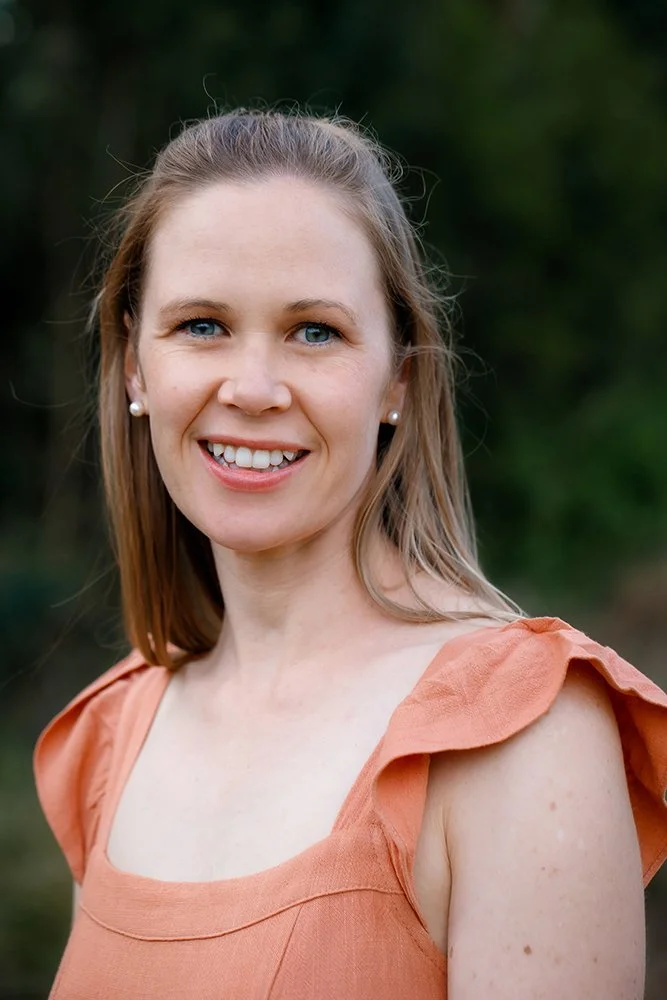 A woman with long blonde hair,blue eyes, and wearing pearl earrings, smiling at the camera outdoors, dressed in an orange sleeveless top with ruffled shoulder details.