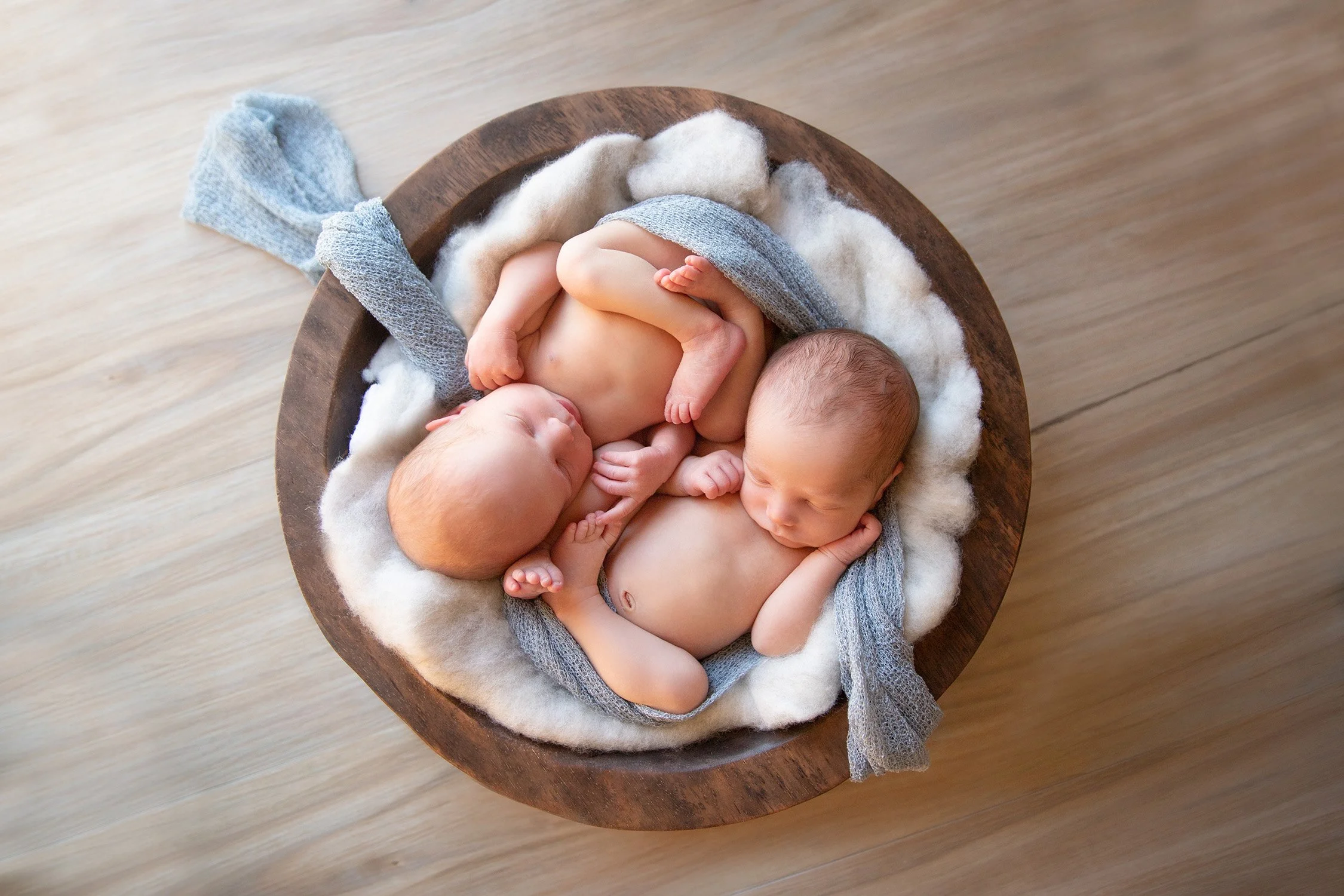 Two sleeping newborn babies curled up together in a round wooden bowl, surrounded by soft white wool and gray knit blankets.