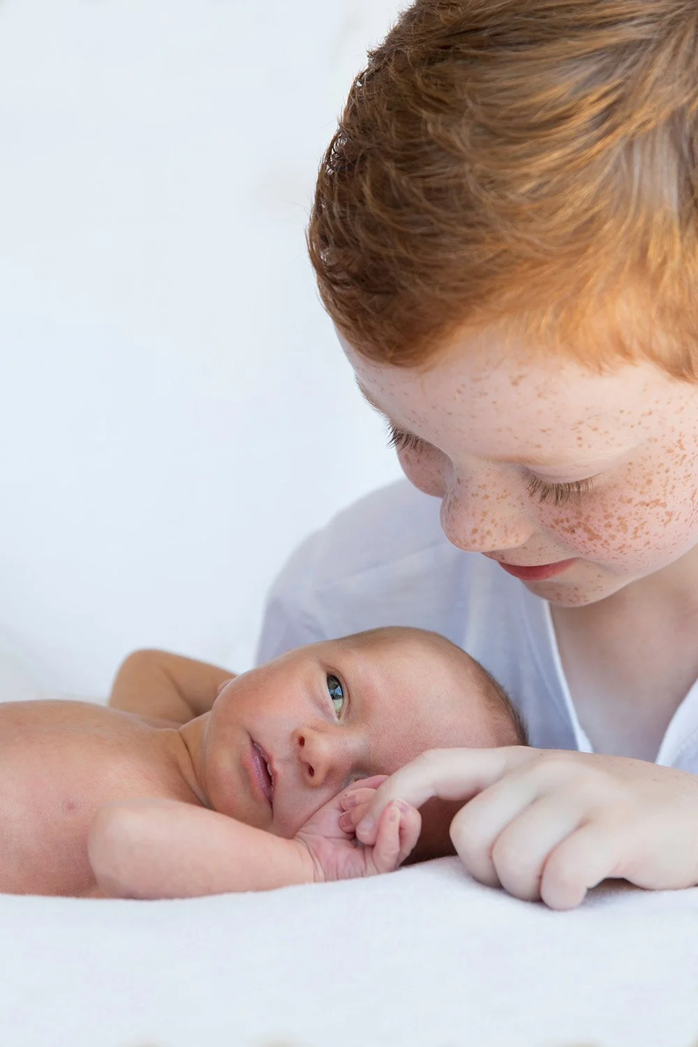 A young red-haired child with freckles looking at a newborn baby with light hair, who is lying down and gazing back.