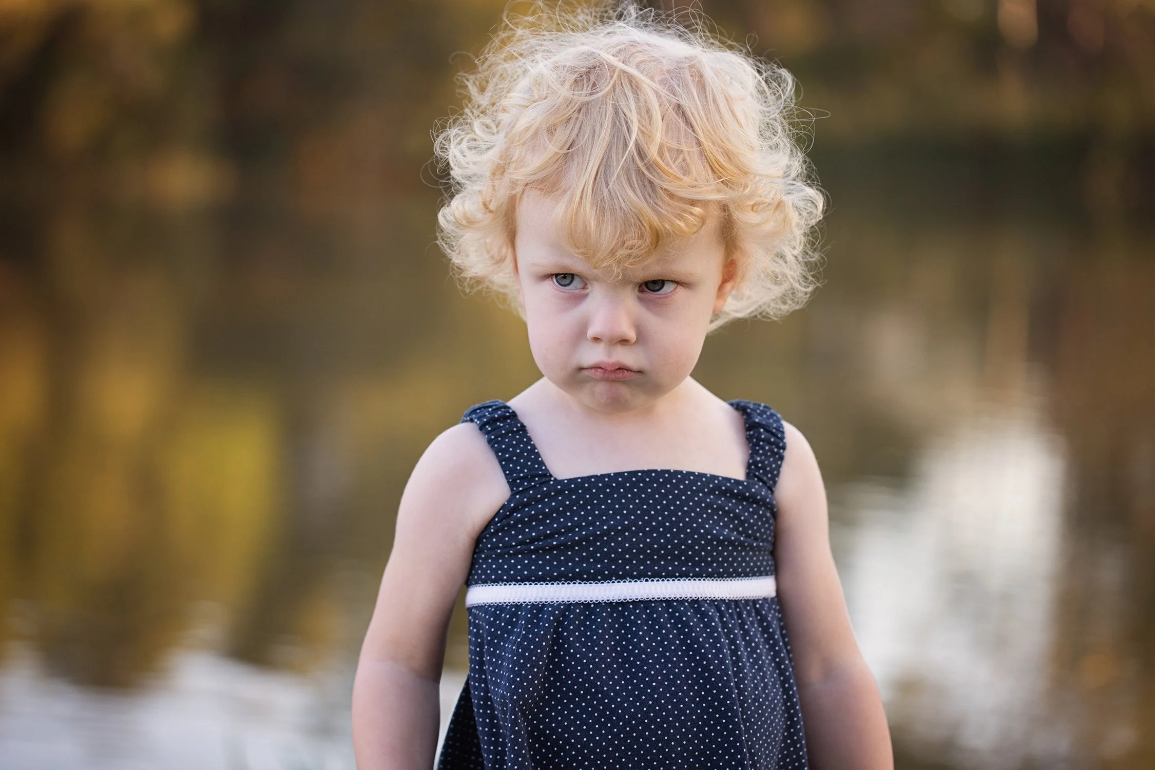 A young girl with curly blonde hair and blue eyes looks sternly, wearing a navy blue dress with white polka dots, outdoors near a body of water.