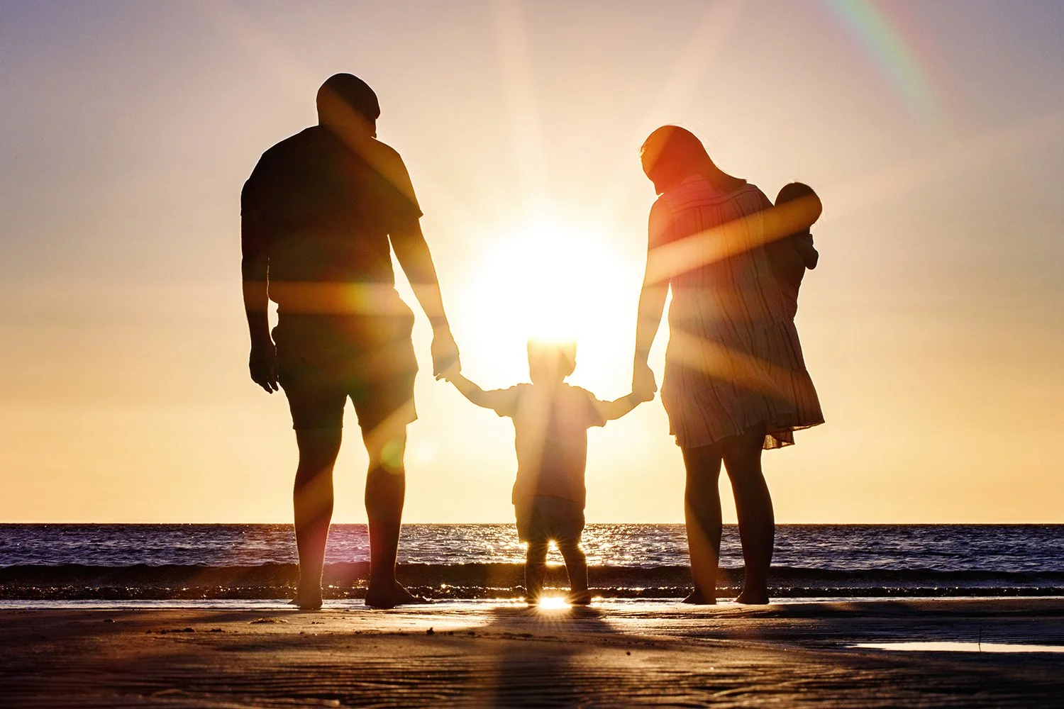 Silhouette of a family of four holding hands on the beach during sunset, facing the ocean.