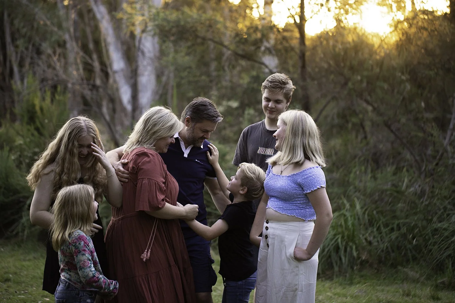 A family of seven, including children and adults, gathering outdoors in a wooded area during sunset, sharing a joyful moment with smiles and affectionate touches.
