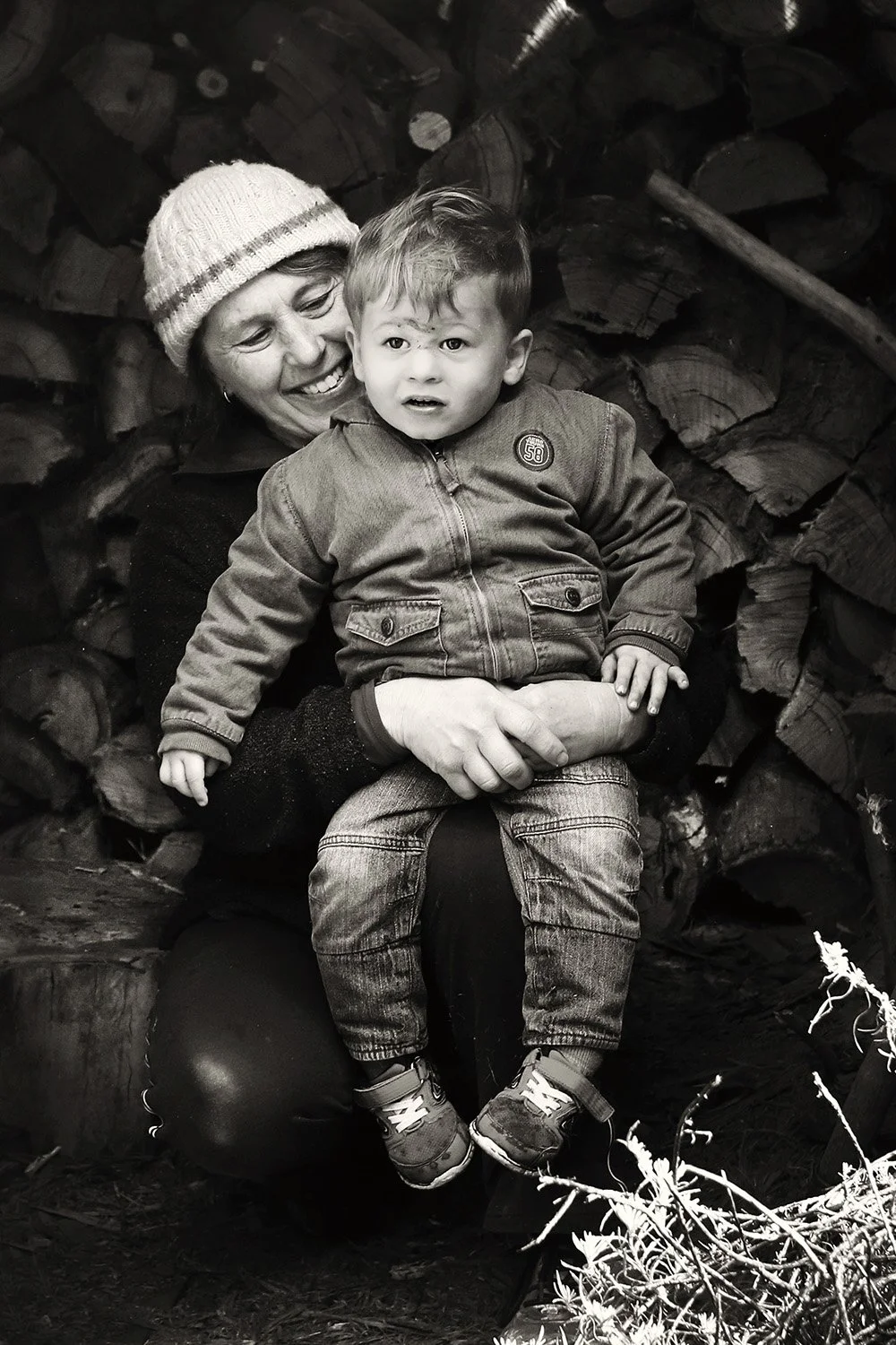 A woman holding a young boy on her lap in front of a background of stacked firewood. The woman is smiling and wearing a knit hat, while the boy looks confused or worried.