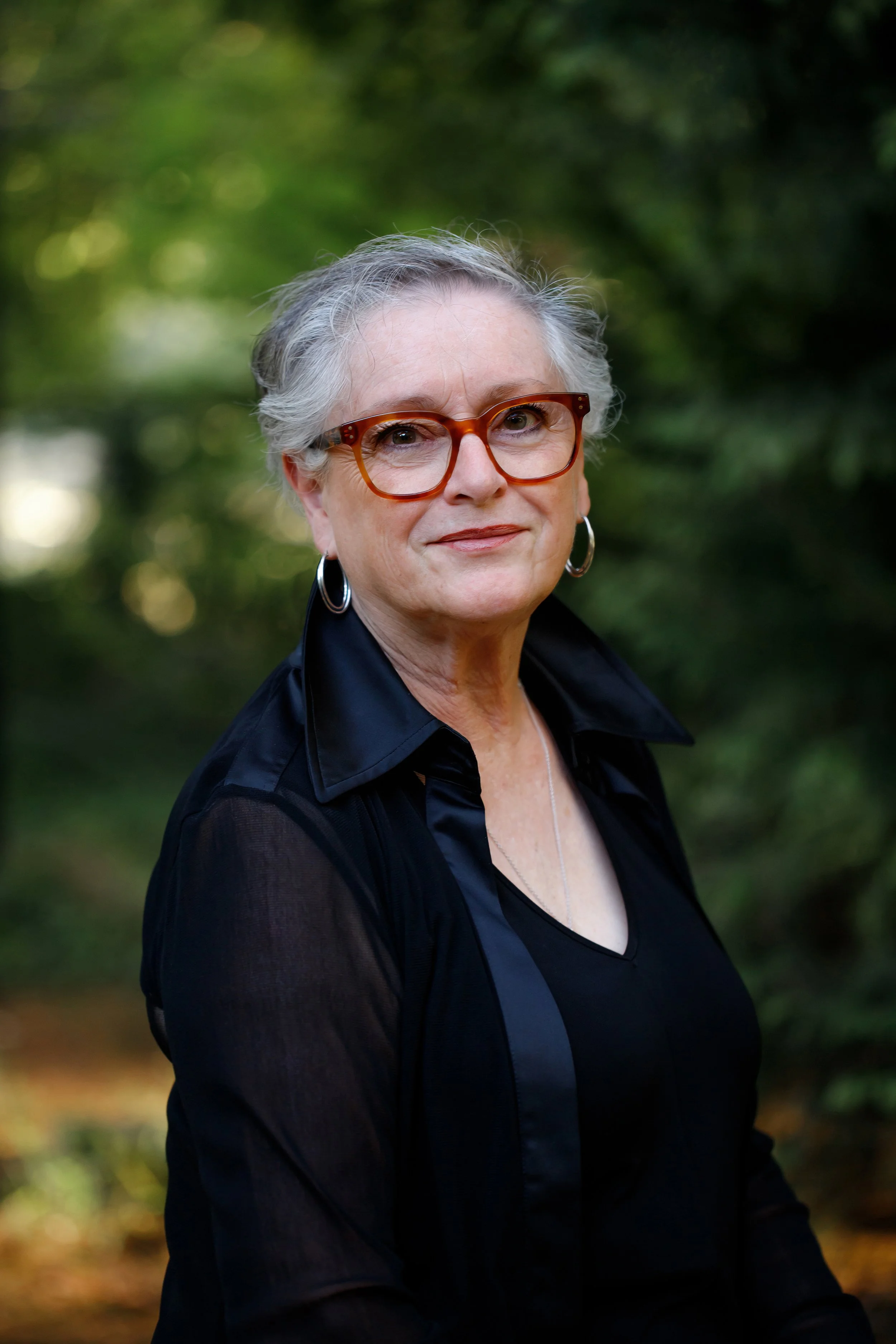 A portrait of an older woman with gray hair, wearing glasses, hoop earrings, a black shirt, and a black jacket, outdoors with green foliage in the background.