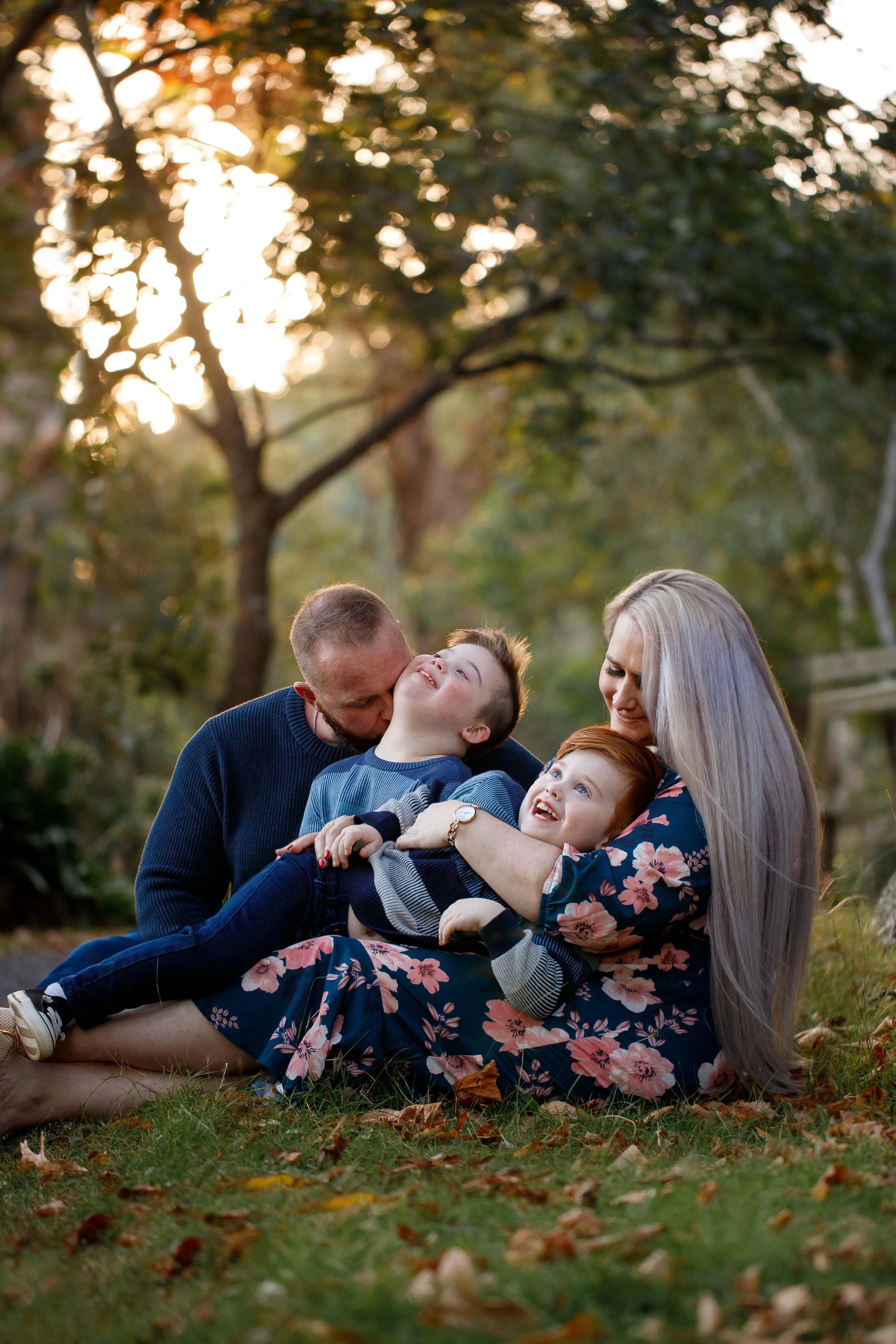 A family of four, including a mother, father, and two children, playing and laughing outdoors on a grassy area with fallen leaves in the fall, with sunlight filtering through trees in the background.