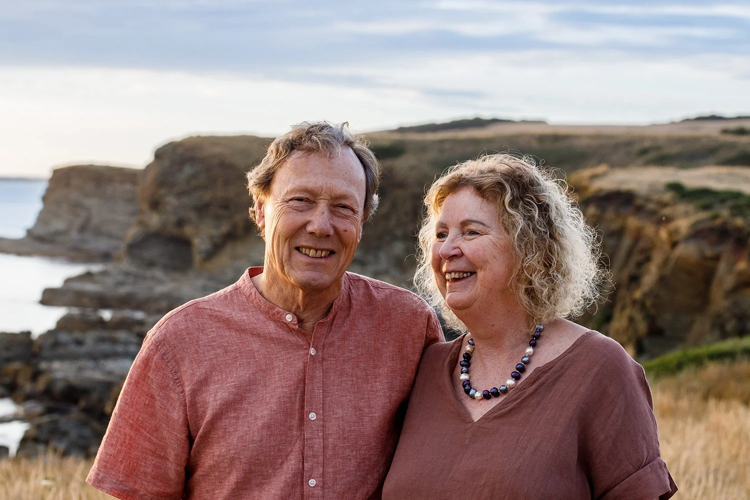 An elderly couple smiling and standing close together outdoors near rocky cliffs with grass and a cloudy sky in the background.