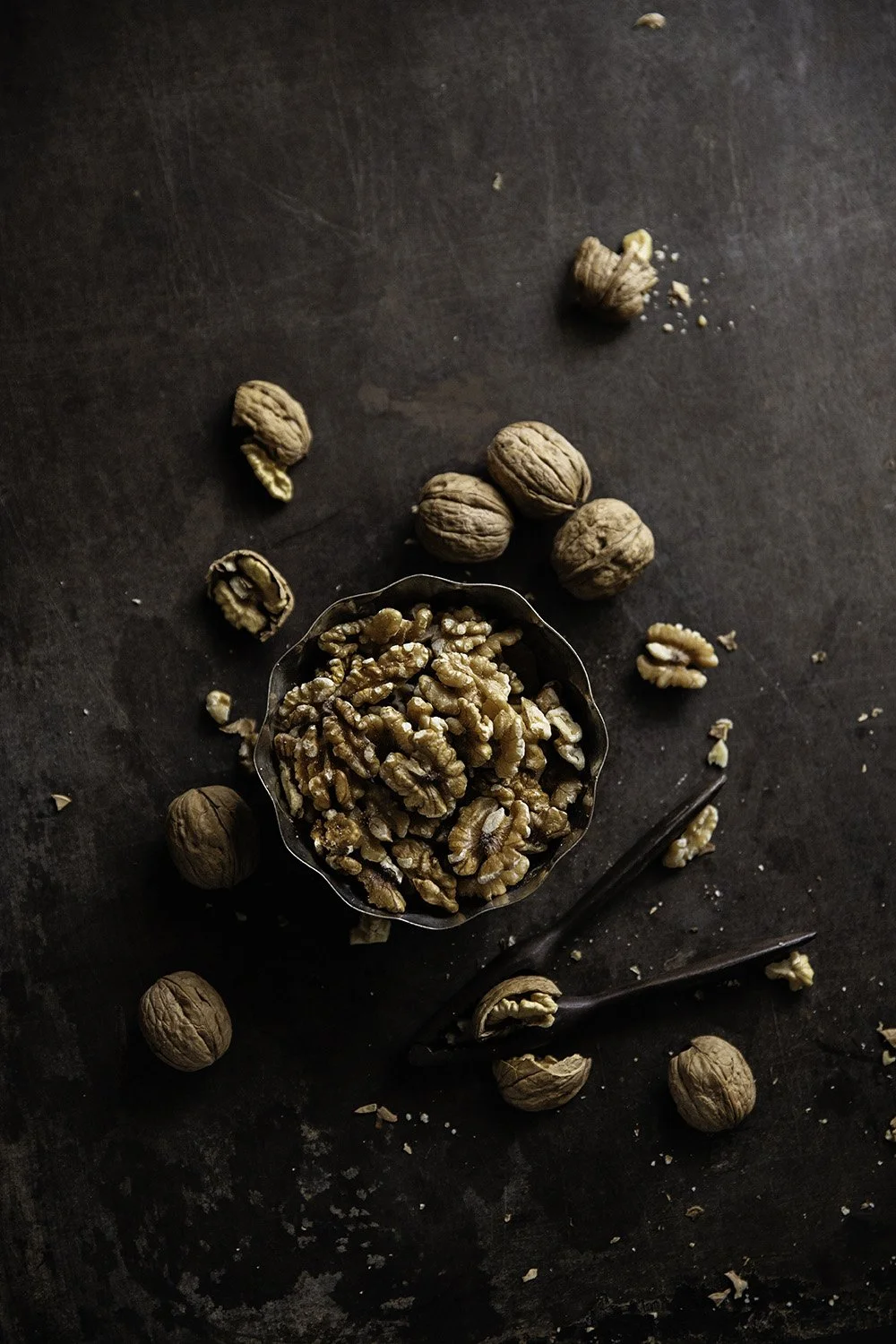 Walnuts and walnut halves on a dark surface, with some in a small metal bowl and others scattered around. Two nutcracker tools are also present.