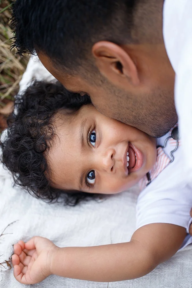 A person with short dark hair and a beard is lying on a white blanket, kissing the cheek of a smiling young child with curly black hair and bright blue eyes.