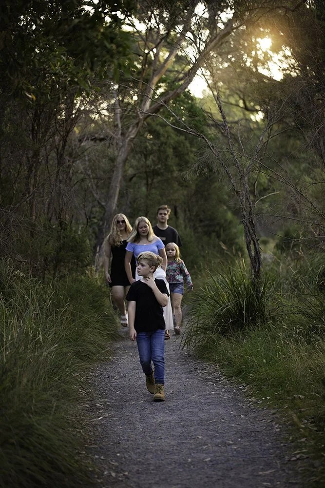 A group of five children and teenagers walking along a dirt trail in a forest at sunset.