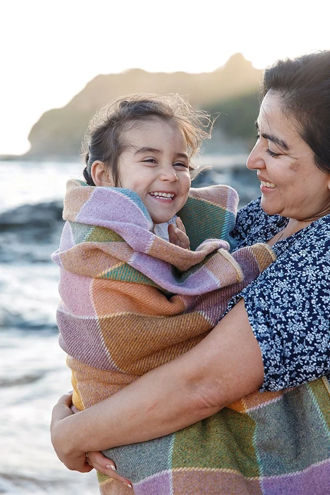 A woman holding a smiling young girl wrapped in a colorful plaid blanket at the beach, with ocean waves and a distant mountain in the background.