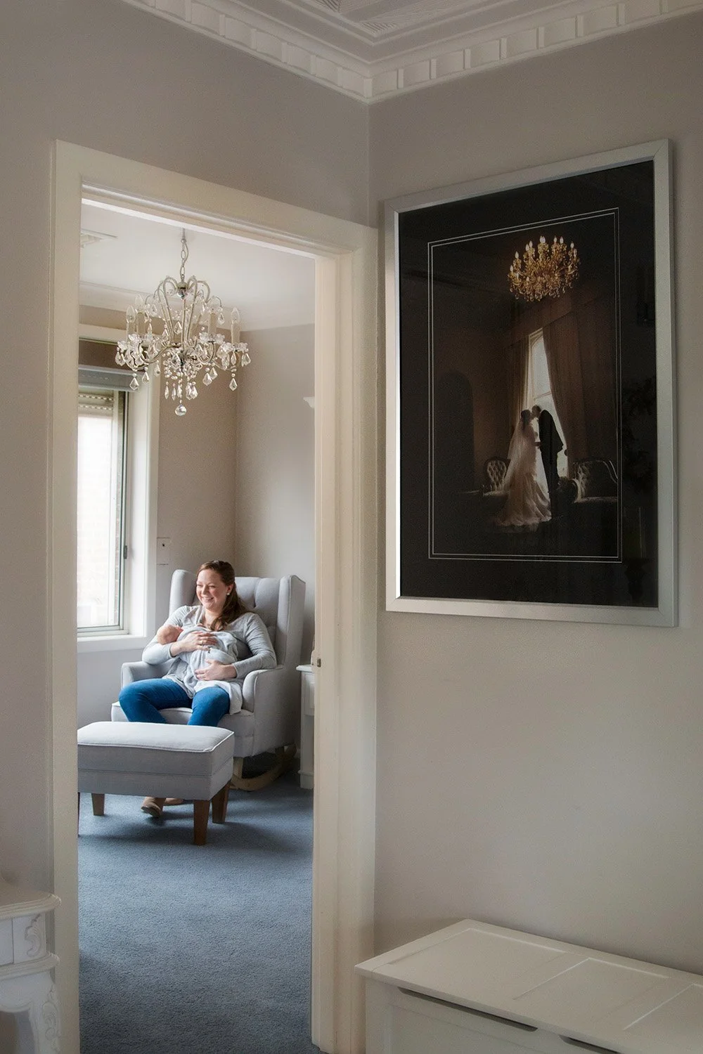 A woman sitting in a plush armchair, smiling and holding her pregnant belly, in a well-lit room with large windows, a chandelier, and a framed wedding photo on the wall.