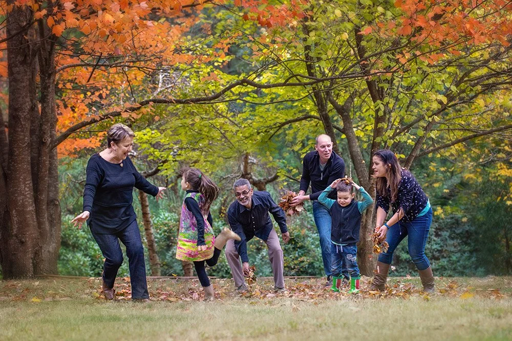 A family of six playing and throwing leaves in a park during fall, with colorful autumn trees in the background.